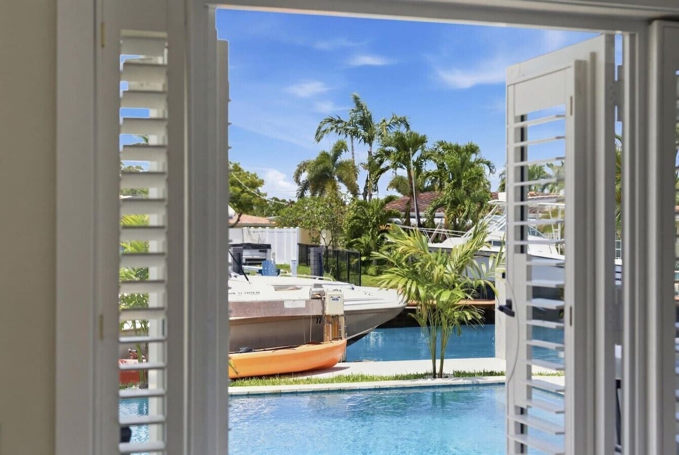 A view through a window of a swimming pool with boats, palm trees, and a clear sky.