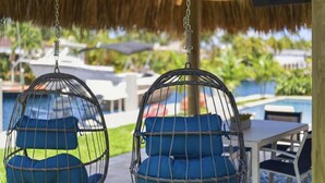 Duas cadeiras suspensas com almofadas azuis, uma piscina e uma estrutura de telhado de palha.