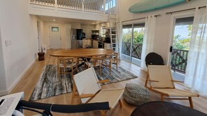 Un vaste espace de vie comprenant une table à manger, des chaises, un balcon avec une planche de surf, et une cuisine en arrière-plan.