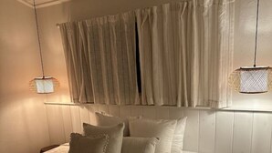 Une chambre avec un grand lit, deux lampes suspendues, une fenêtre avec des rideaux et un tabouret en bois.