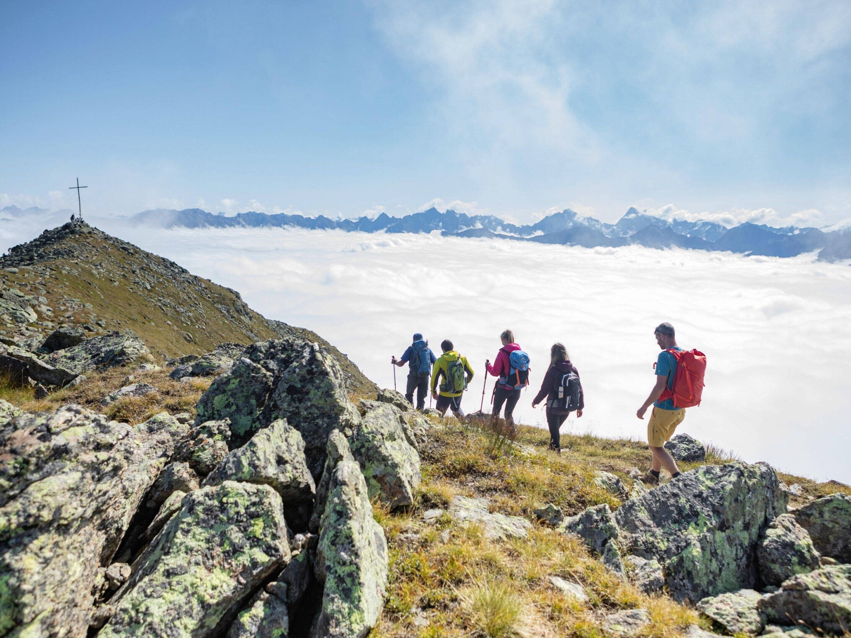Un grupo de excursionistas en un sendero de montaña con terreno rocoso y una cruz a lo lejos.