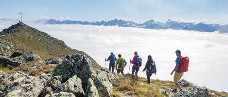 Un grupo de excursionistas en un sendero de montaña con terreno rocoso y una cruz a lo lejos.