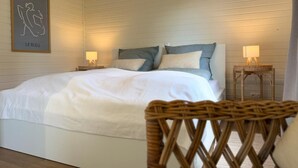 Een slaapkamer met een bed, twee bedlampen, een rieten stoel en een ingelijst schilderij aan de muur.