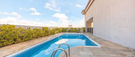 A rectangular swimming pool with clear blue water, surrounded by stone tiles and a low stone wall.