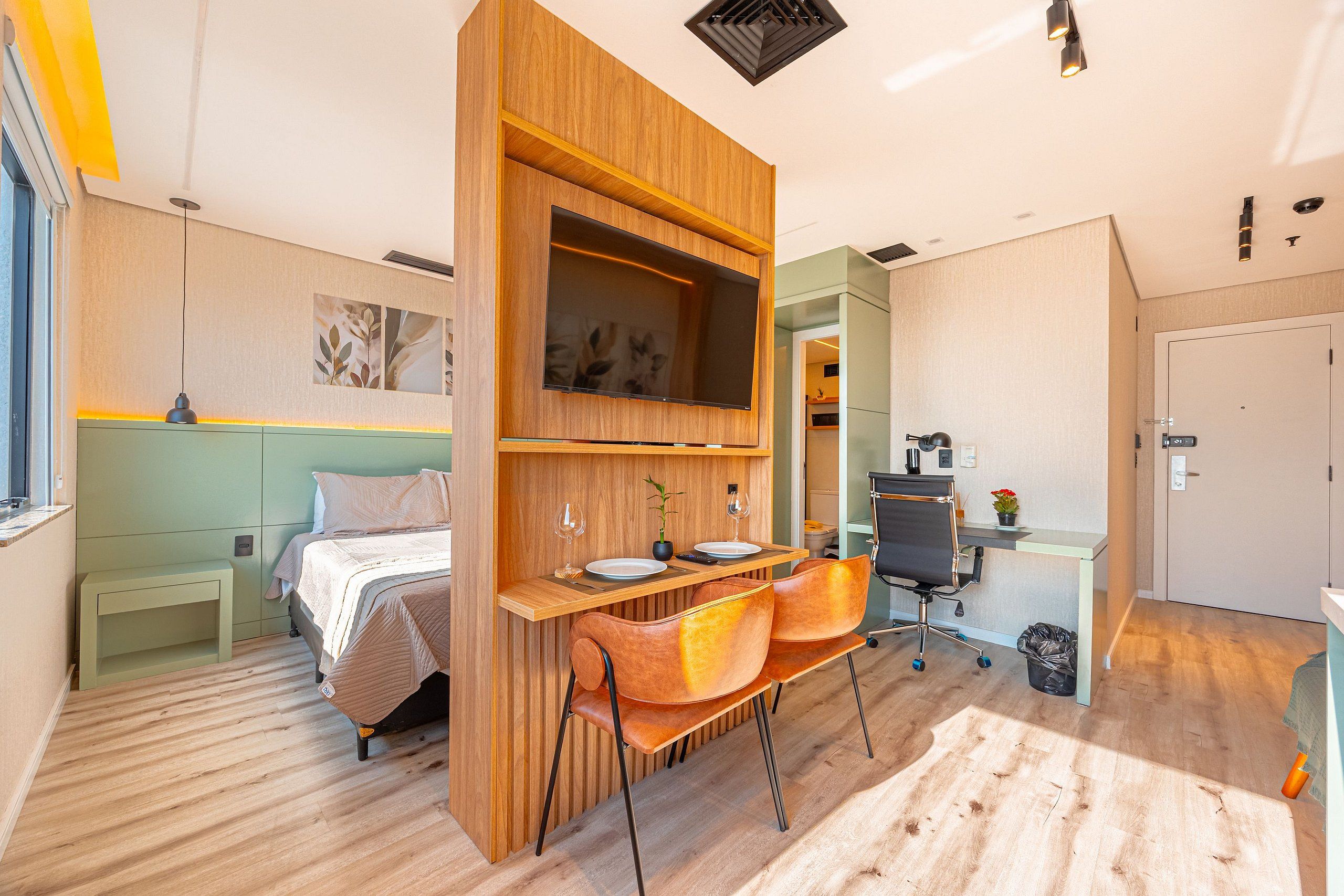 A compact hotel room with a bed, a wooden desk, two chairs, a TV, and a small table with a plant.