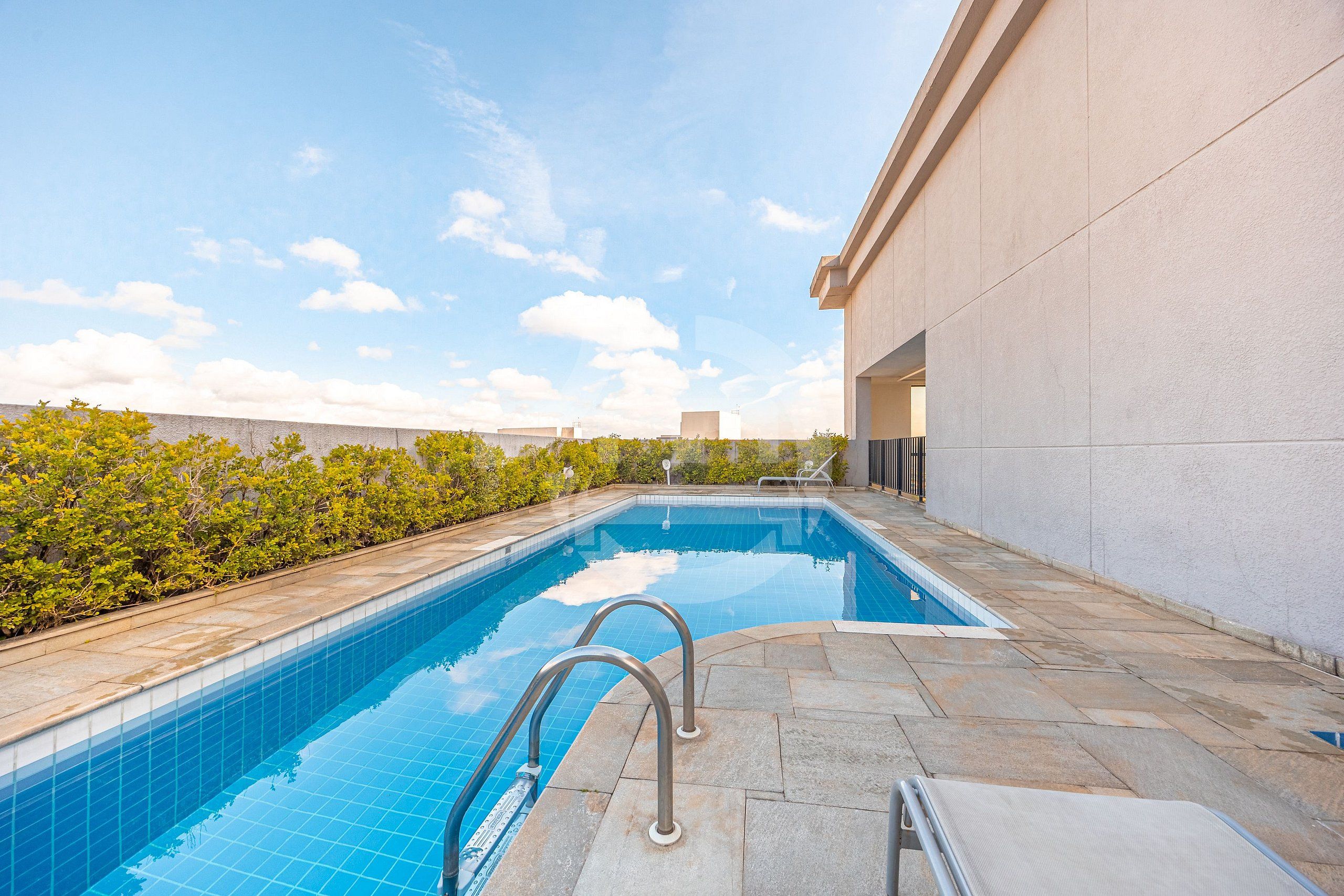 A rectangular swimming pool with clear blue water, surrounded by stone tiles and a low stone wall.