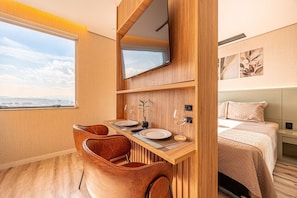 Quarto moderno com escrivaninha de madeira, duas cadeiras, uma cama e uma televisão fixada na parede.