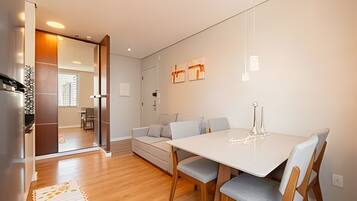 A modern living space with a dining area, a sofa, and a kitchen visible through an open door.