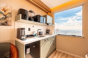 Cozinha compacta com máquina de lavar louça, cafeteira, micro-ondas e liquidificador na bancada. Há uma janela com vista para a cidade.