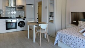 Ein kompaktes Hotelzimmer mit Küchenzeile, einem Bett mit floralem Überwurf und einer kleinen Essecke mit Tisch und Stühlen.