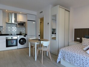 Ein kompaktes Hotelzimmer mit Küchenzeile, einem Bett mit floralem Überwurf und einer kleinen Essecke mit Tisch und Stühlen.