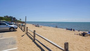 Une plage avec une clôture en bois, des voitures garées et des gens qui se détendent au bord de l’eau.