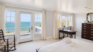 A bedroom with a bed, a desk, and a dresser. There are large windows with curtains and a view of the ocean. - Private Beach, Sweeping Water Views, Game Room! (Brewster)