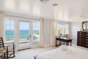 A bedroom with a bed, a desk, and a dresser. There are large windows with curtains and a view of the ocean.