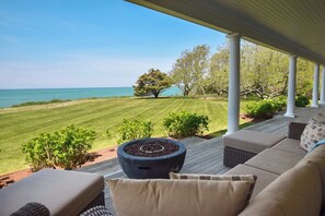 A covered outdoor seating area with a view of the ocean, a fire pit, and a lawn.