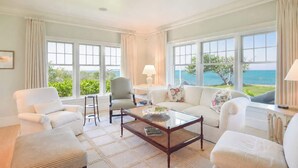 A spacious living room with a large window overlooking a scenic view, a comfortable seating area with a coffee table, and a view of the ocean. - Private Beach, Sweeping Water Views, Game Room! (Brewster)