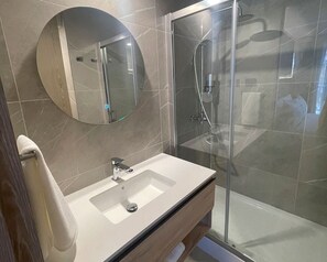 A modern bathroom with a glass-enclosed shower, a round mirror, and a white sink. - One Manquehue EXC | Bright 1BR Apartment (Santiago)