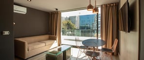 Ein modernes Wohnzimmer mit einer Couch, einem Glastisch, einem Esstisch mit Stühlen und einem großen Fenster mit Vorhängen.