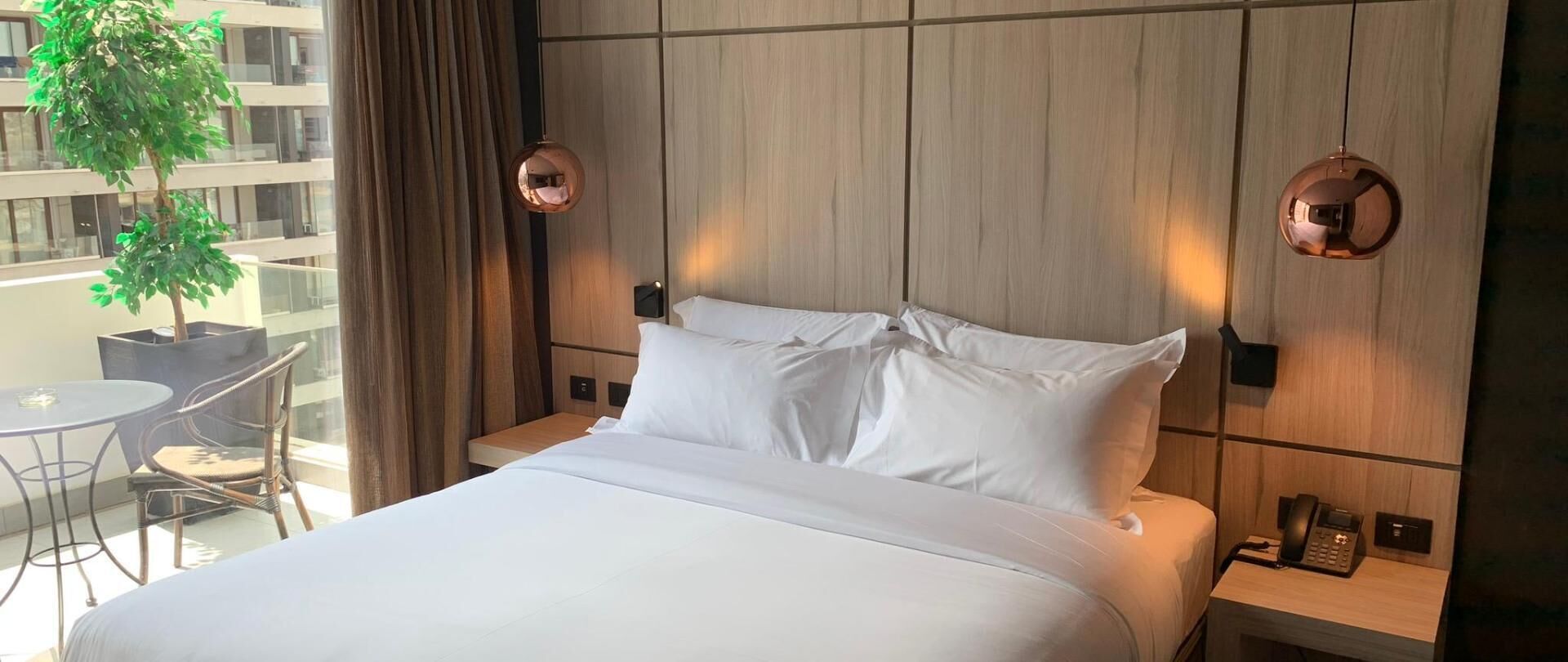 Een hotelkamer met een bed, twee nachtkastjes, een klein tafeltje met stoelen, een potplant en uitzicht op een gebouw.