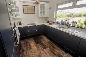 A kitchen with dark cabinets, white countertops, a wooden floor, a window with a view, and a map on the wall. - 4 Cardell Way (Newquay)
