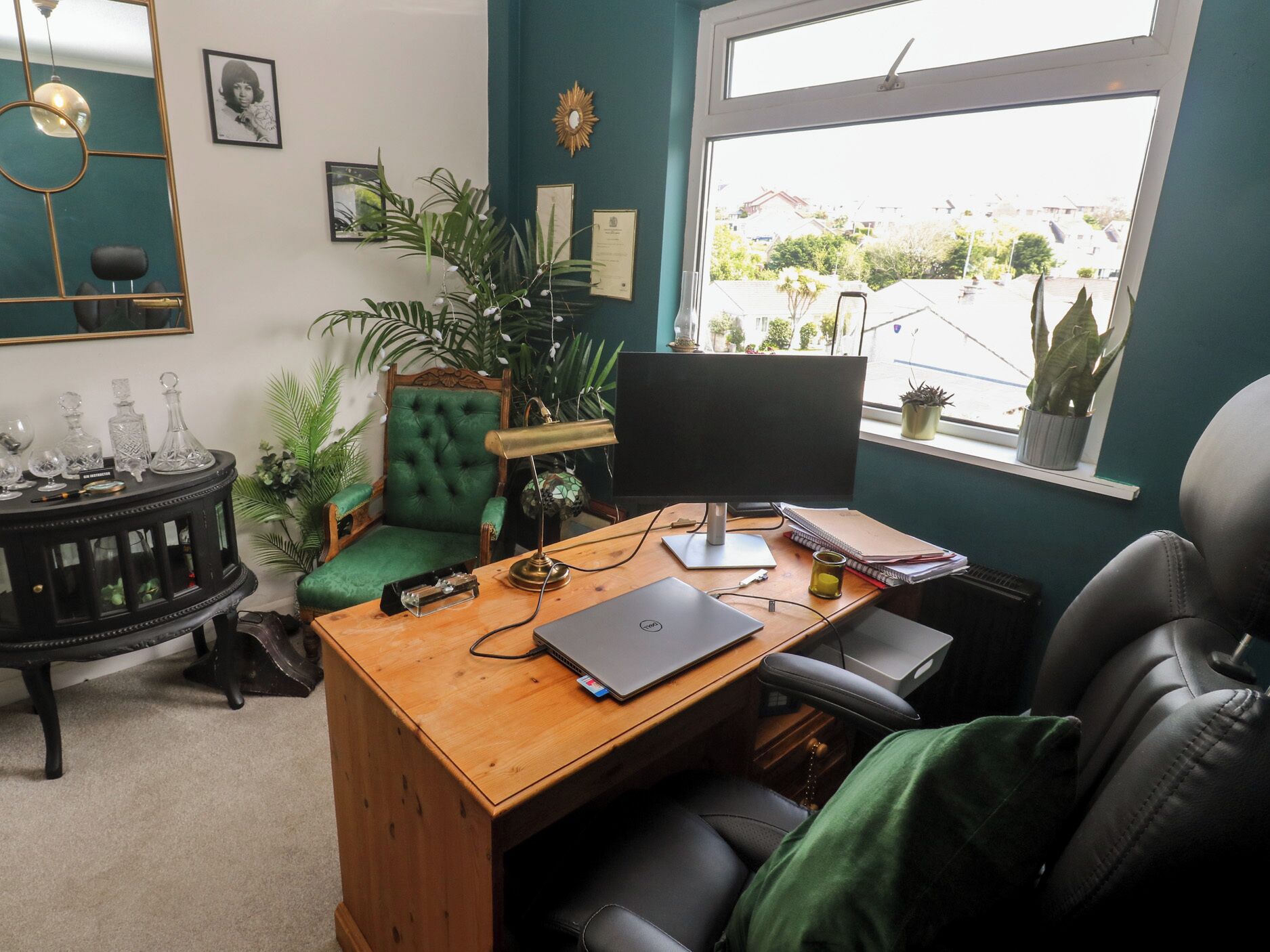 A room with a wooden desk, a computer, a green chair, a black office chair, a lamp, and a window with a view of buildings.