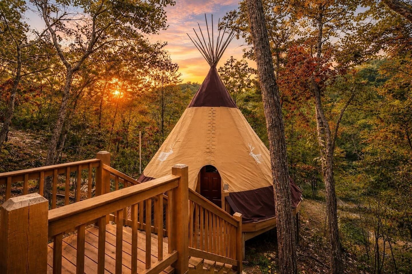A wooden deck leading to a teepee with a deer symbol, surrounded by trees and a sunset.