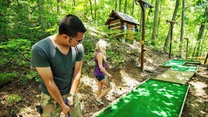 A person playing mini-golf in a wooded area with a small wooden structure in the background.