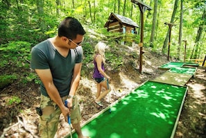 A person playing mini-golf in a wooded area with a small wooden structure in the background.