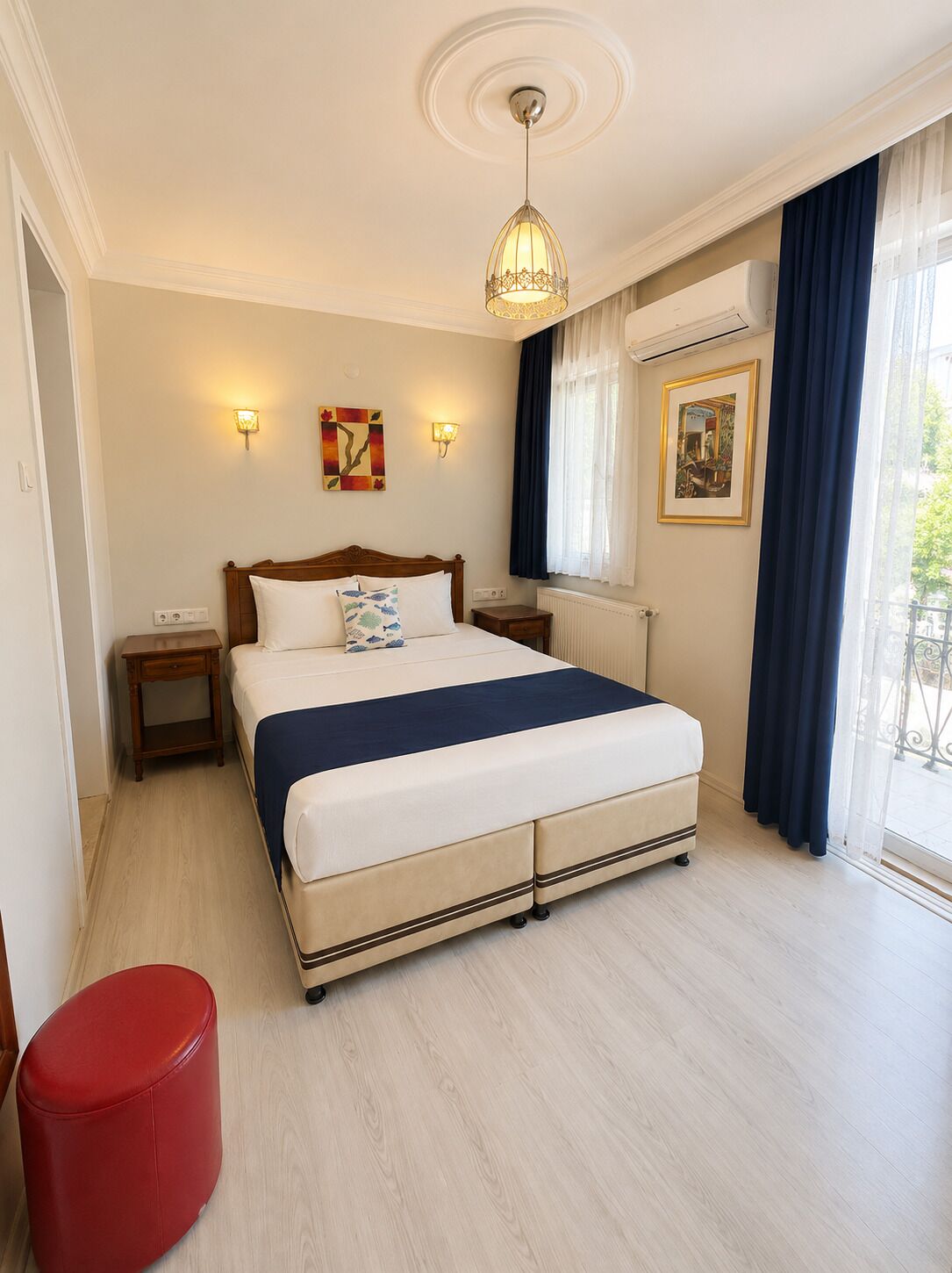 Hotelkamer met een bed, nachtkastjes, een rode poef, een plafondlamp en een raam met gordijnen.