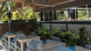 Un patio al aire libre con mesas blancas y sillas azules, faroles colgantes y plantas en macetas.