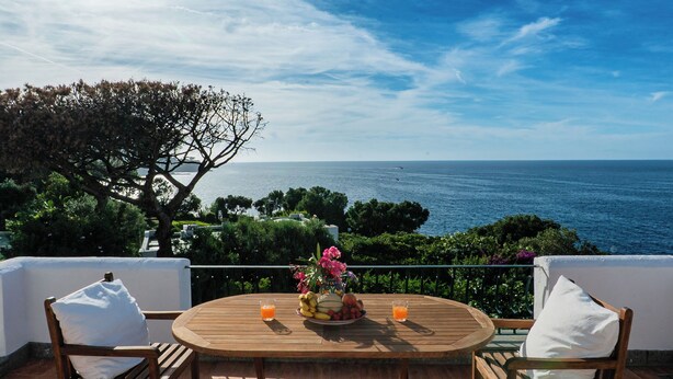 A round wooden table with a fruit plate and two glasses of orange juice, overlooking a coastal view with a clear sky. - Bouganville by IschiaExp (Ischia Porto)