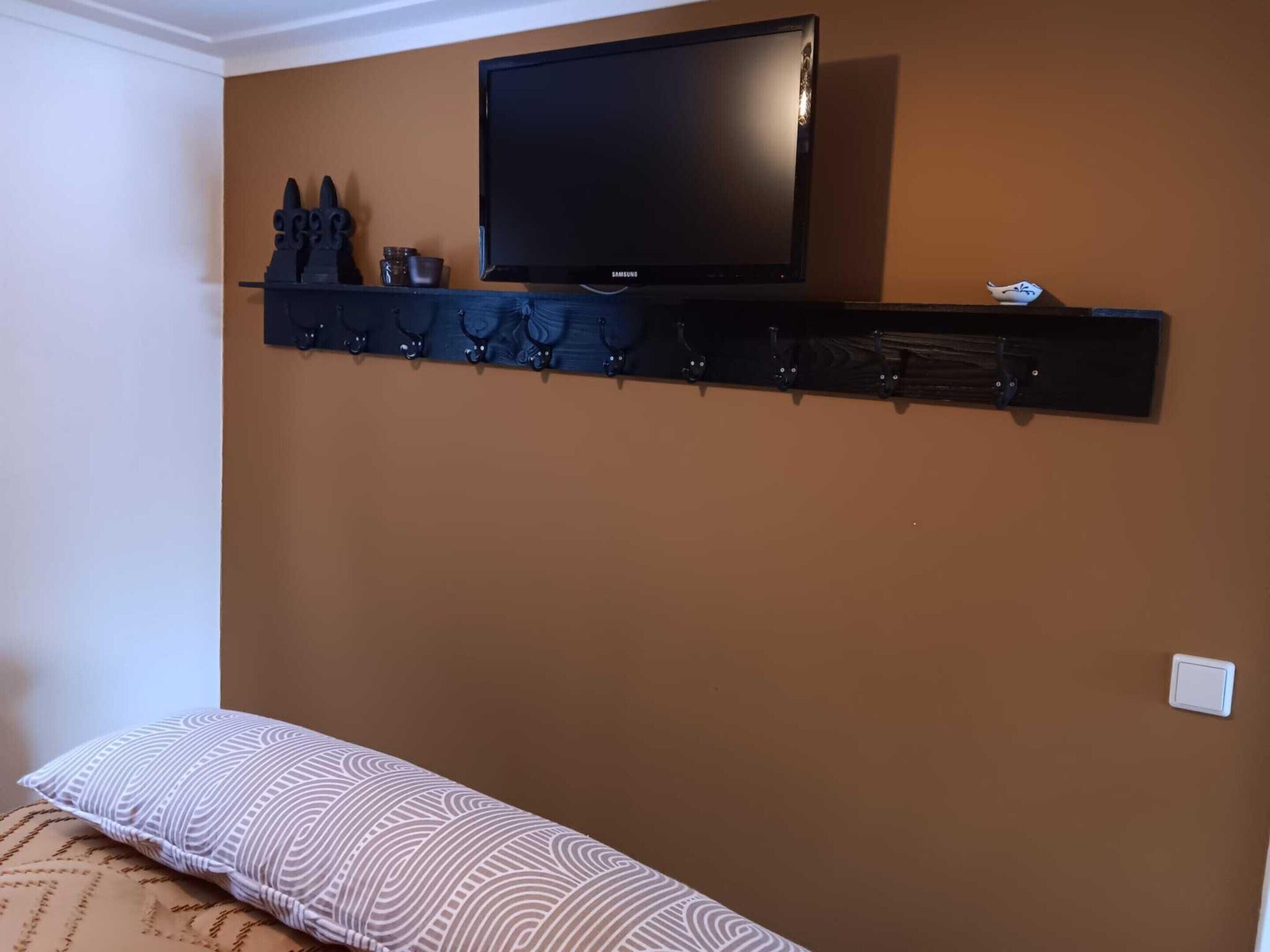 A room with a brown wall, a mounted TV, a black shelf with hooks, and a bed with patterned bedding.