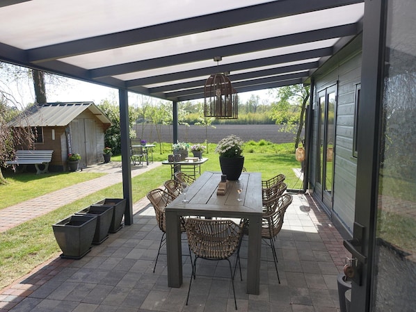 A covered patio with a table and chairs, potted plants, and a view of the surrounding garden.