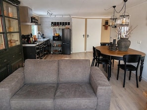 A modern kitchen with a built-in oven, a dining area with a wooden table and chairs, and a grey sofa in the living room.