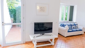 Uma sala de estar com uma TV sobre uma mesa branca, um sofá branco com almofadas azuis e um quadro na parede.