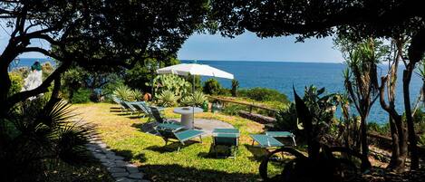 A garden with a stone pathway leading to a seating area with chairs and a table, overlooking a body of water.