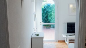 Um corredor com piso ladrilhado, uma porta branca que dá para o exterior e uma pequena mesa com uma planta.