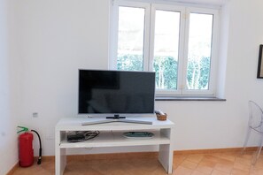 Una habitación con televisión sobre un pedestal blanco, un extintor y una ventana con vista a árboles.