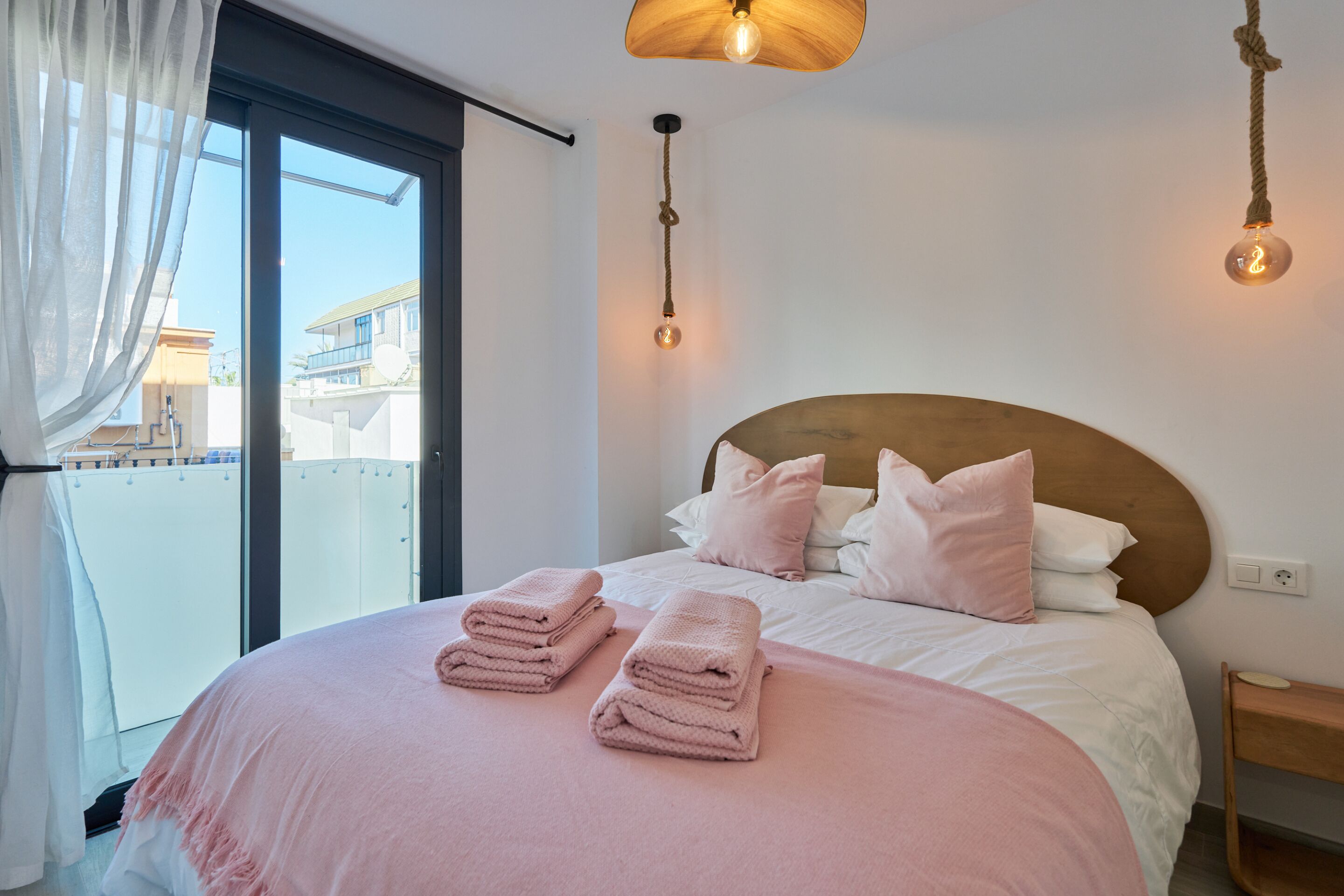 Un lit bien fait, agrémenté de serviettes roses, avec une tête de lit en bois, et une vue sur l’extérieur grâce à une grande fenêtre.