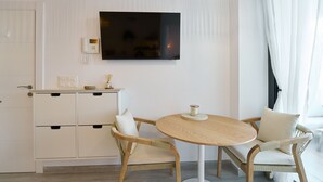 Ein modernes Zimmer mit Flachbildfernseher an der Wand, einem kleinen Holztisch mit Kerze, zwei Korbstühlen und einem weißen Schrank mit Schubladen.