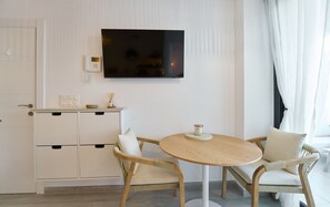 Ein modernes Zimmer mit Flachbildfernseher an der Wand, einem kleinen Holztisch mit Kerze, zwei Korbstühlen und einem weißen Schrank mit Schubladen.