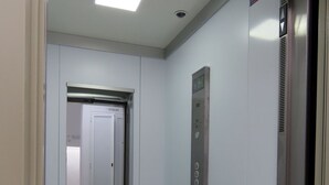 Ein schmaler Korridor mit einer Tür, einem Wandbedienfeld für den Aufzug und einer Deckenleuchte.