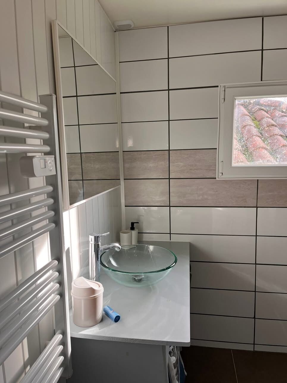 Ett badrum med ett handfat i glas, vita kakelplattor, en handdukshängare och ett fönster med utsikt.