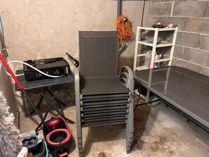 Een kamer met een stoel met metalen frame, een tafeltje met elektronische apparatuur, een bezem en een plank met diverse spullen.