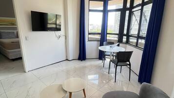 Uma sala de estar moderna com sofá, duas mesinhas de centro, televisão e uma janela grande com cortinas.