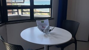 Ein Zimmer mit einem runden Tisch, zwei Stühlen, einem Fenster mit Blick auf Strand und Gebäude sowie einem Metallgestell mit einer Wasserflasche.