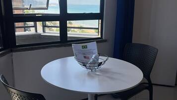 Quarto com mesa redonda, duas cadeiras, janela com vista para a praia e edifícios, e um suporte de metal com uma garrafa de água.
