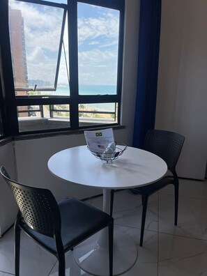 Ein Zimmer mit einem runden Tisch, zwei Stühlen, einem Fenster mit Blick auf Strand und Gebäude sowie einem Metallgestell mit einer Wasserflasche.
