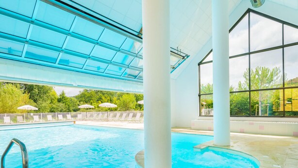 En indendørs swimmingpool med glasloft, hvide søjler og udsigt til grønne områder.