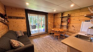 Interior de una cabaña de madera con sofá, mesa de comedor, sillas, una zona de cocina con lavaplatos y un ventanal corredizo que da a un patio exterior.